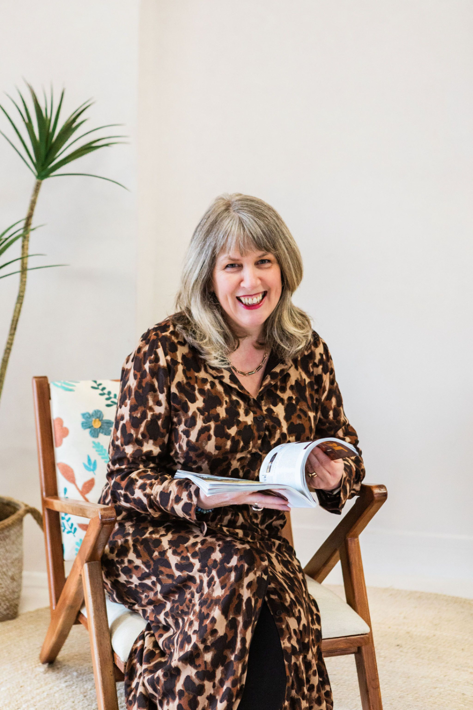 Woman wearing a black long sleeved T shirt and cream trousers, smiling while sitting cross legged on a cream sofa with a stripey throw in neutral tones