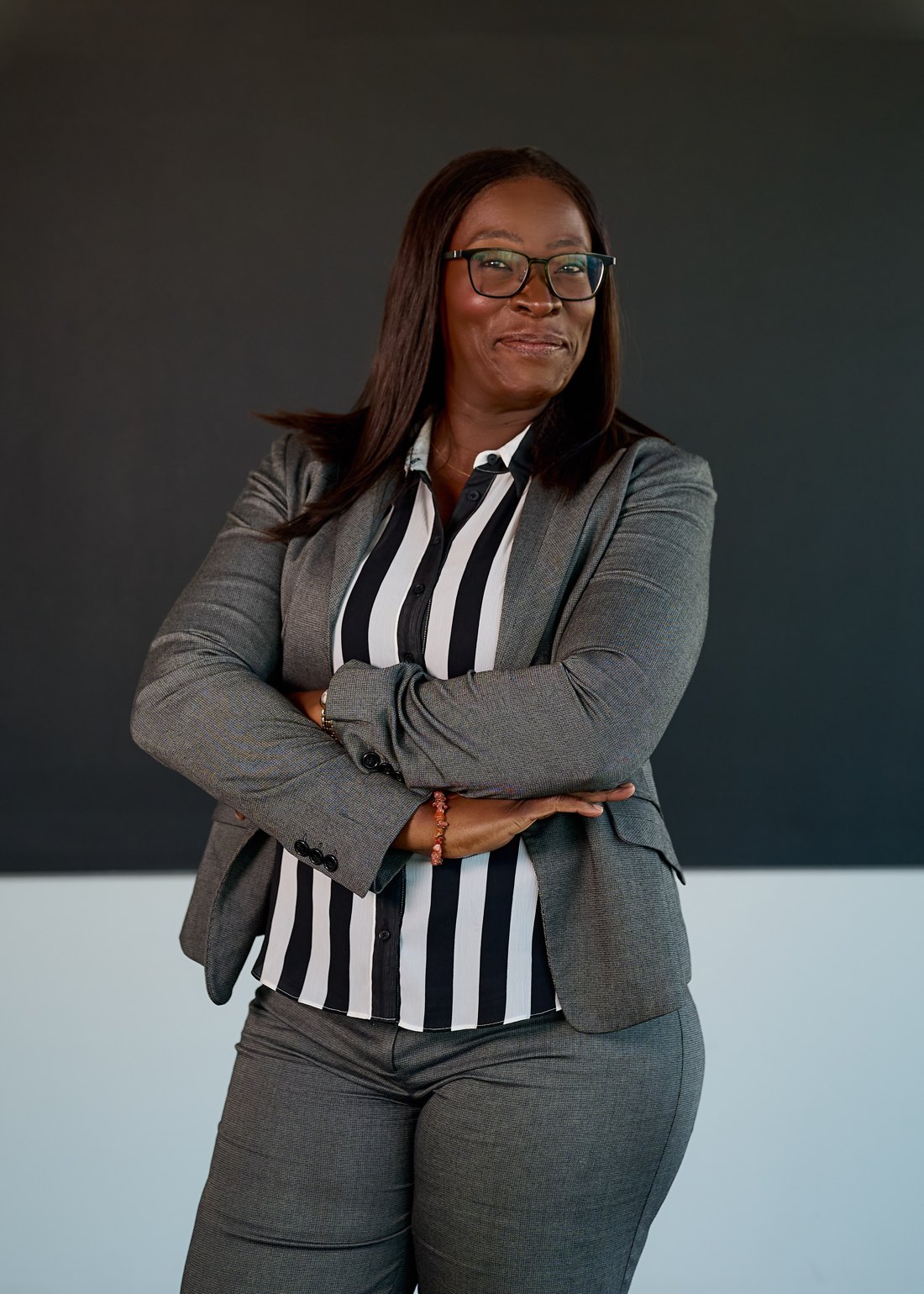 Dr. Shade Okedara standing with her arms folded, wearing glasses and a grey suit