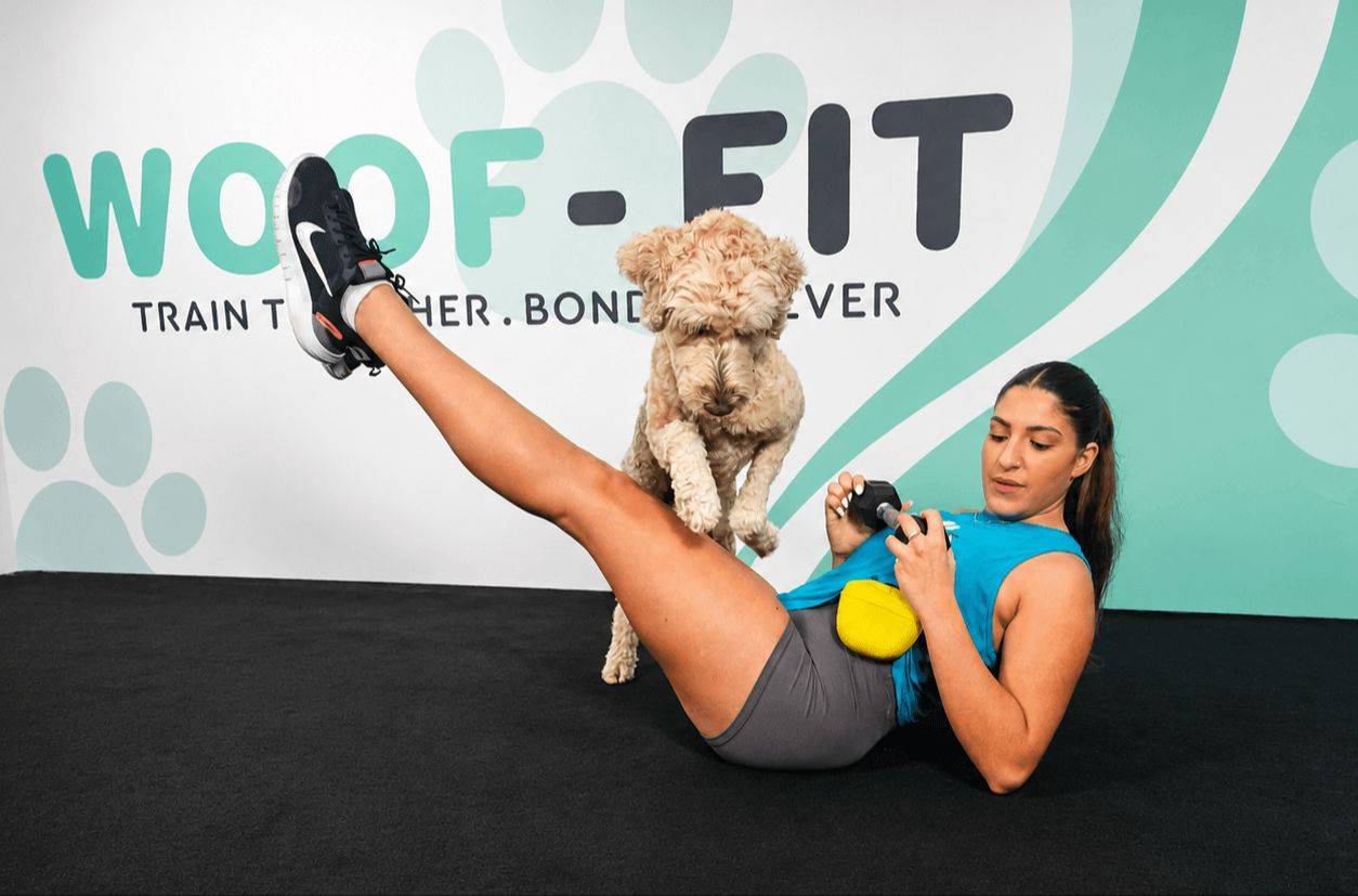 Woman performing a leg raise exercise while her dog jumps over her legs during a synchronized Woof-Fit workout