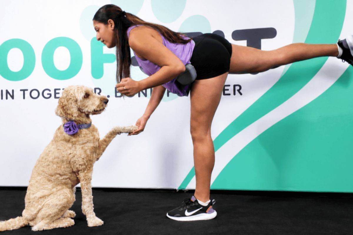 Woman doing a single-leg deadlift exercise while rewarding her Goldendoodle with a treat during the Legs & Paws workout