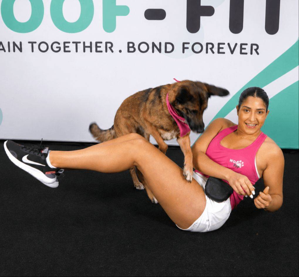 Woman doing a Russian-twist exercise while rewarding her Goldendoodle for jumping over her legs with a treat during a Woof-Fit workout
