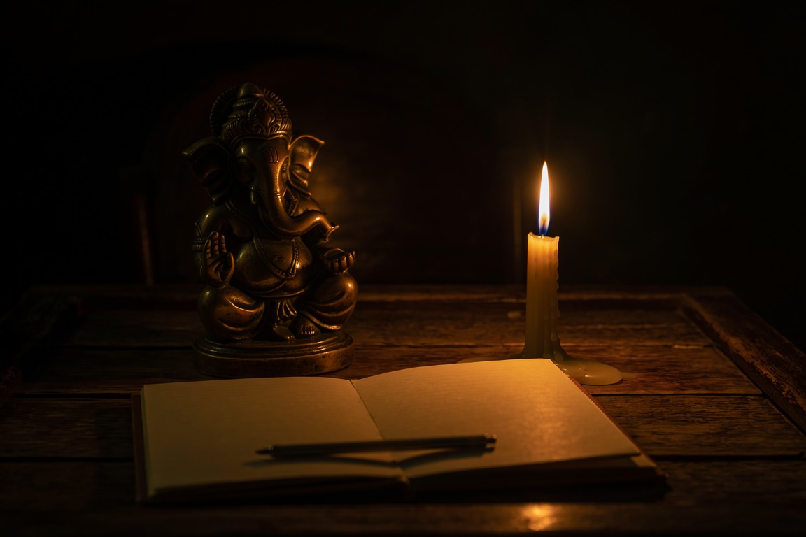 A small bronze figure on a desk beside a lit candle and an open notebook