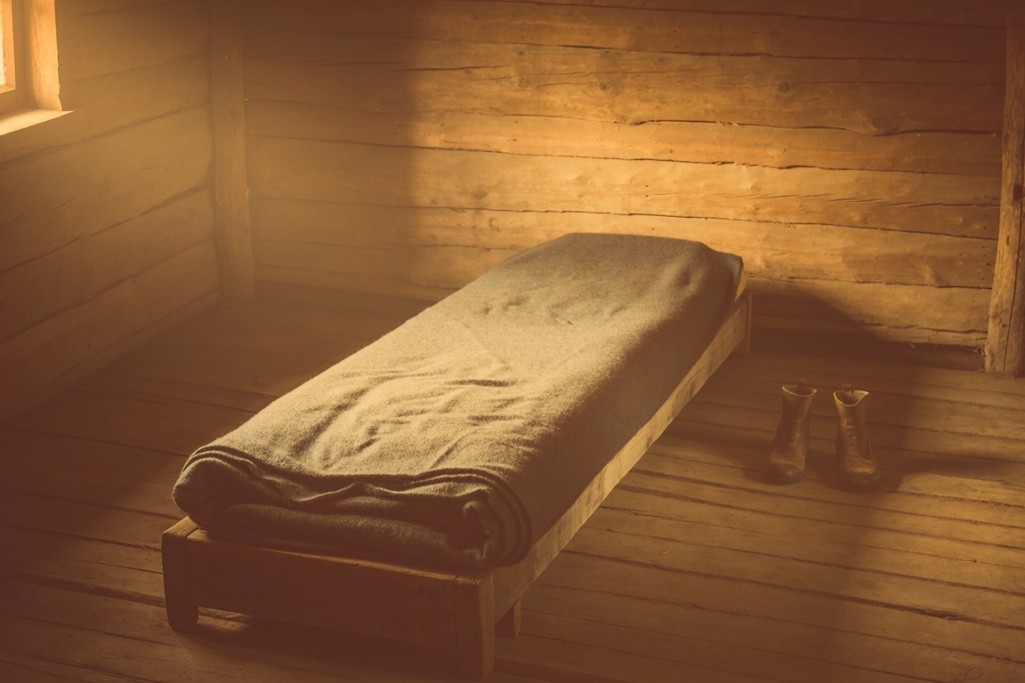 A neatly made narrow bed in a bare timber room, a pair of shoes beside it
