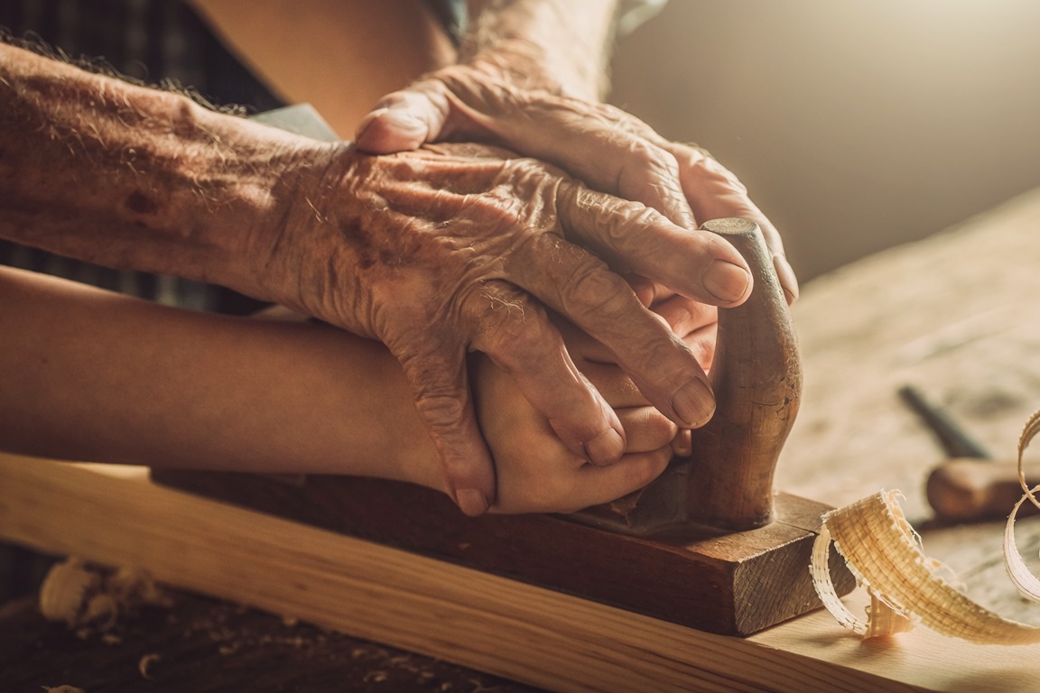 An old craftsman's hands guiding an apprentice's hands on a hand plane over timber