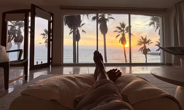 Bare feet resting on a daybed as the sun sets through the windows behind the palms