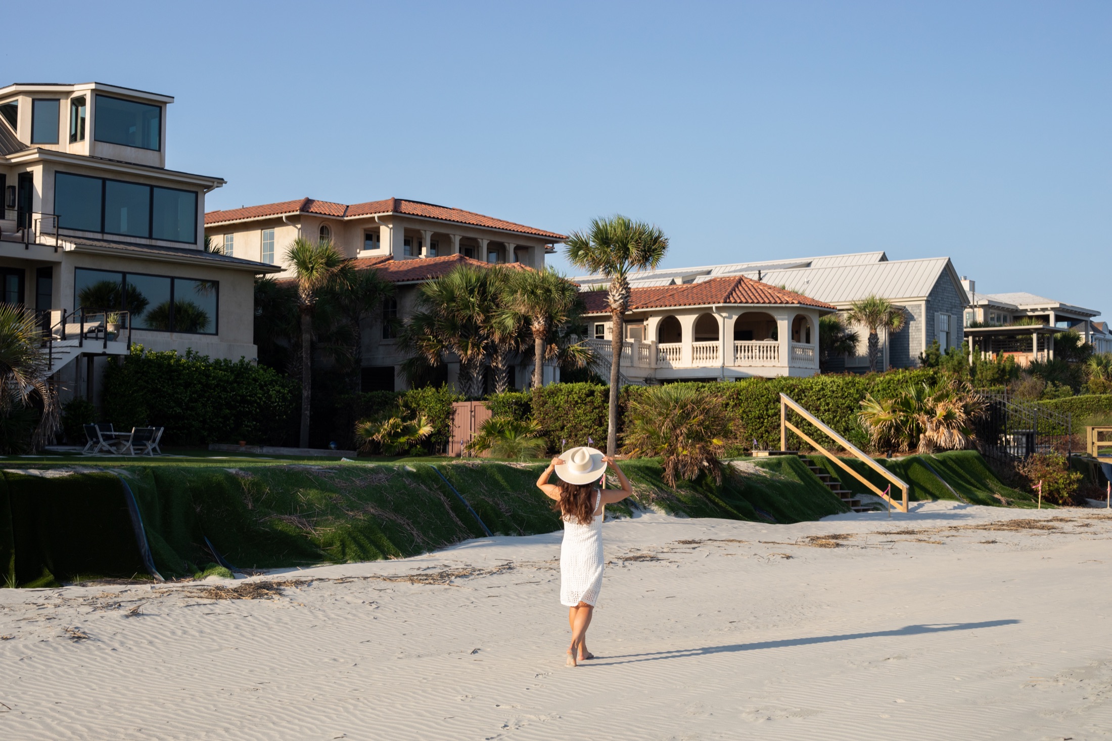 Woman in a white dress and wide-brim hat walking along an empty beach at golden hour, the shoreline stretching ahead of her