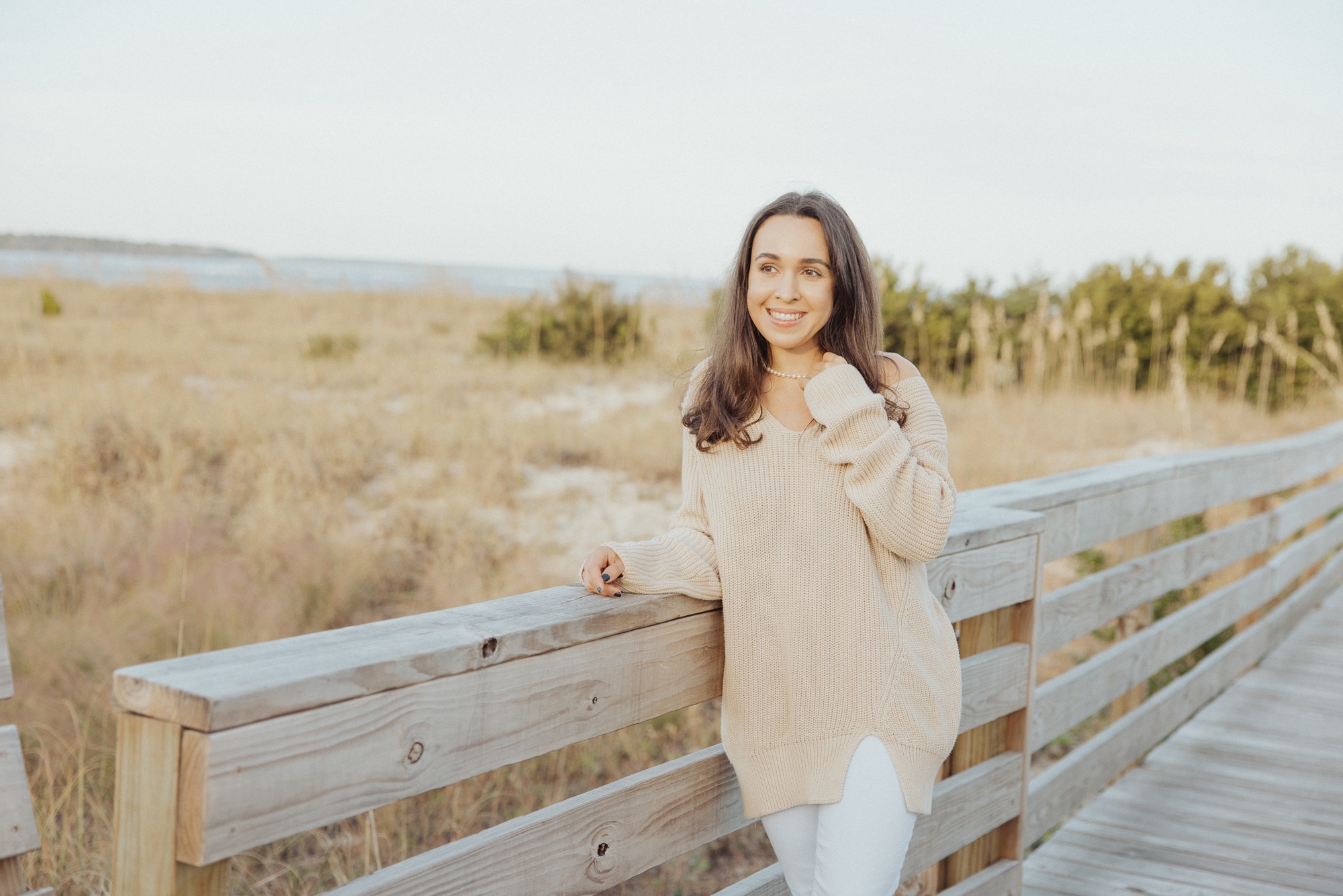 Jacqueline smiling, leaning against a weathered boardwalk railing with dunes, marsh grass and water behind her