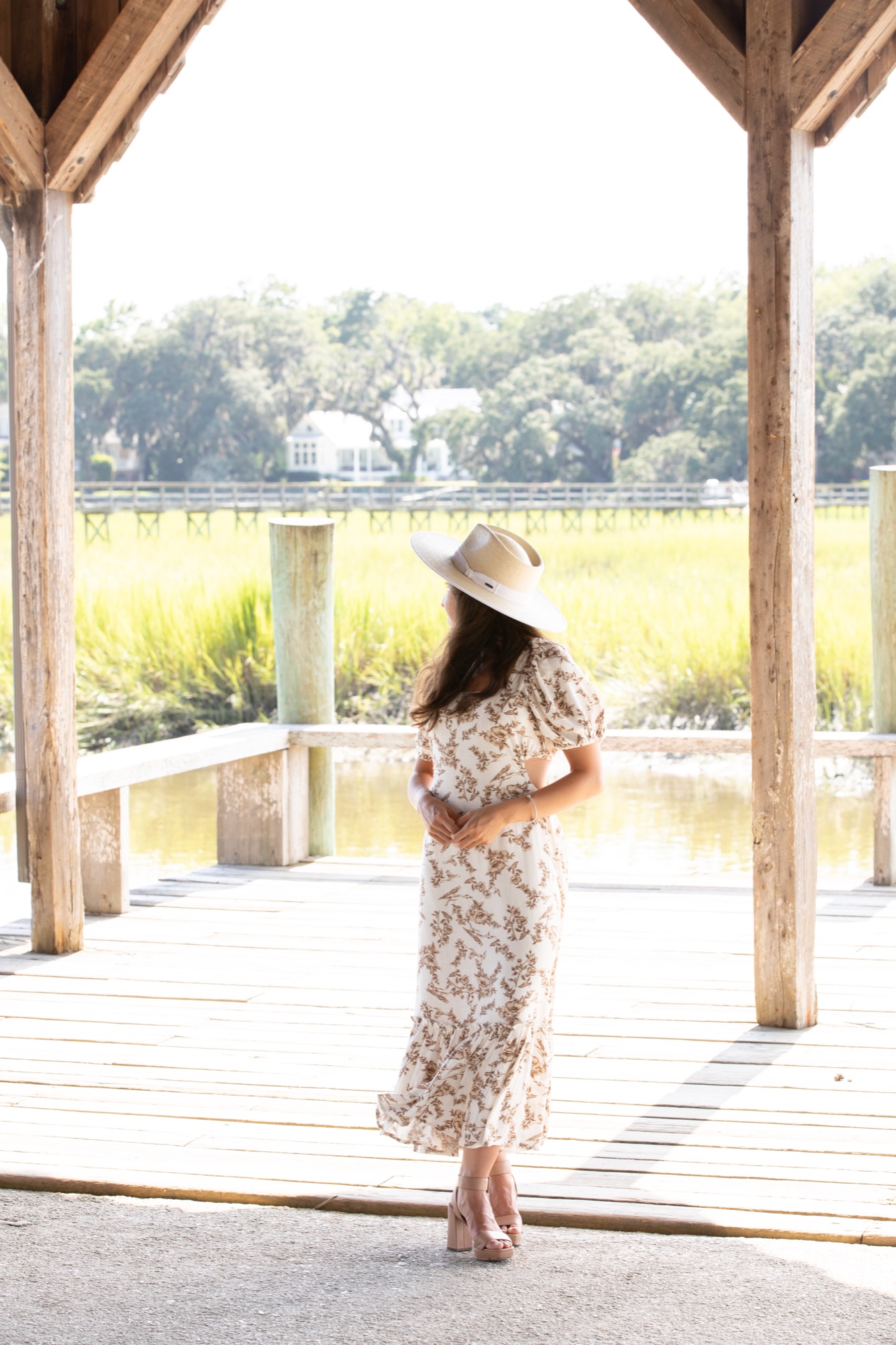 Woman in a floral dress and wide-brim hat standing at the edge of a dock, looking out over marsh grass and open water
