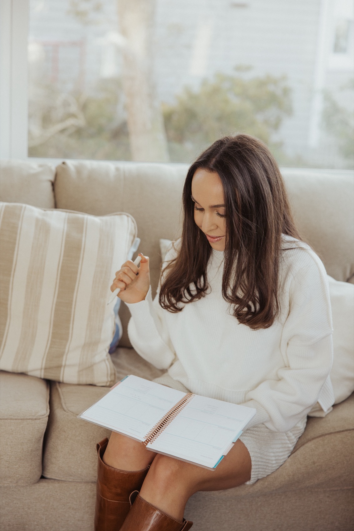 Woman in a cream sweater sitting on a sofa, pen in hand, working through a guided workbook in soft window light
