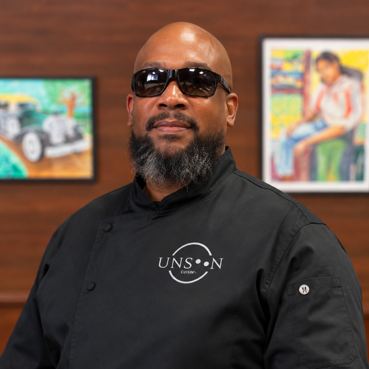 Chef Riq wearing dark glasses and a black Unseen Cuisine® chef coat, standing indoors in front of framed artwork.