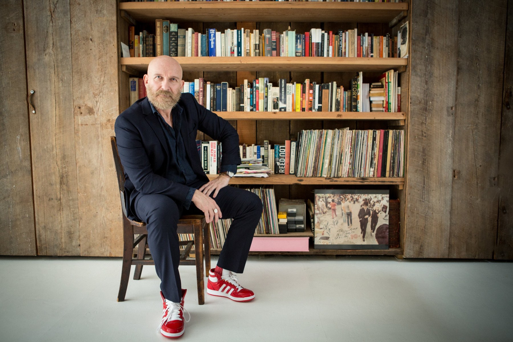 John seated in a dark suit and red sneakers in front of bookshelves
