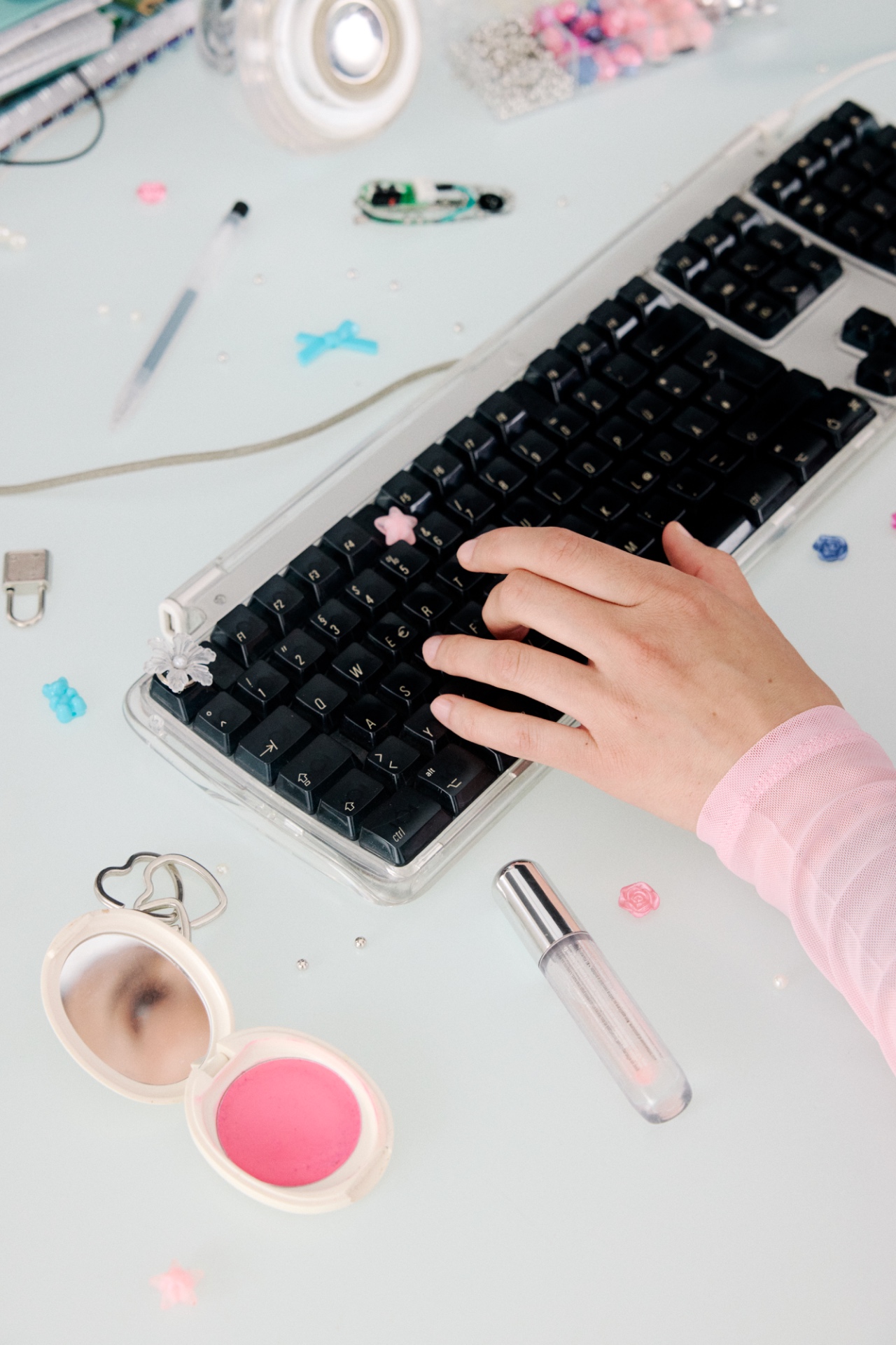 A desk scattered with a keyboard, lipstick, compact, and tech parts
