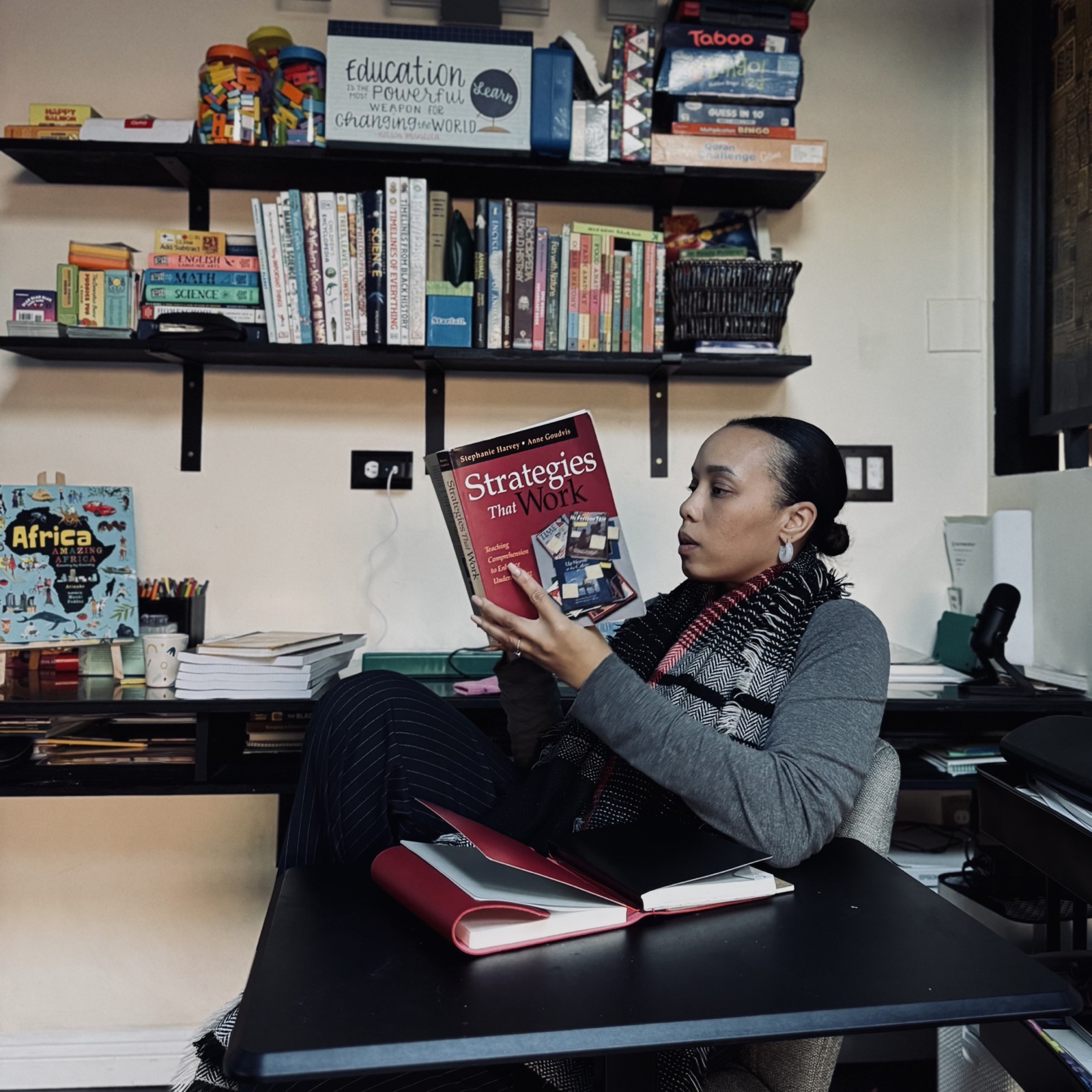 Shahidah at her desk, reading a book with a shelf of books behind her