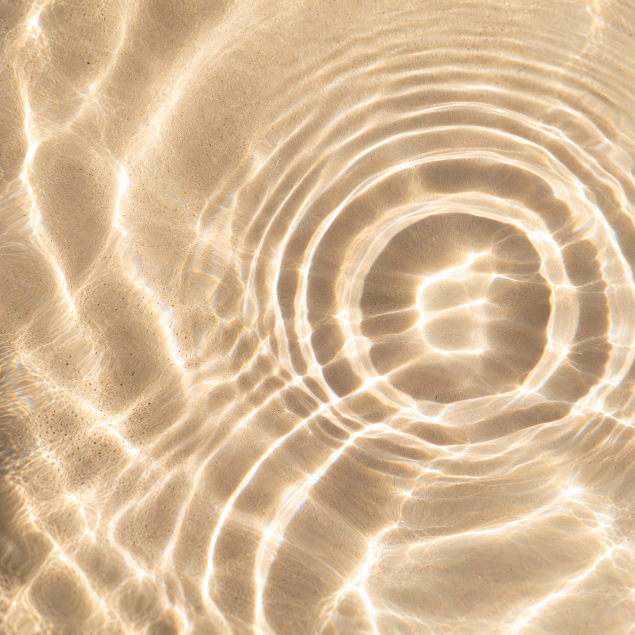 Gentle circular ripples in clear, shallow water over golden sand, with sunlight creating soft, shimmering patterns across the surface.