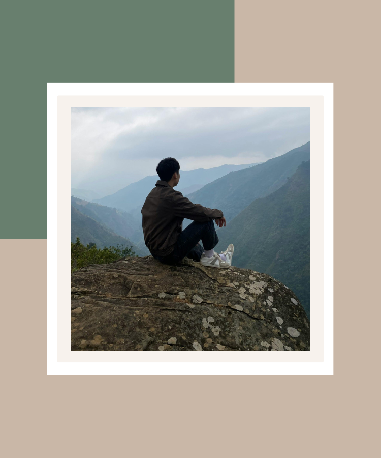 A man sitting on a rocky mountain overlook, gazing towards misty blue mountains, framed by a minimalist sage green and beige background.