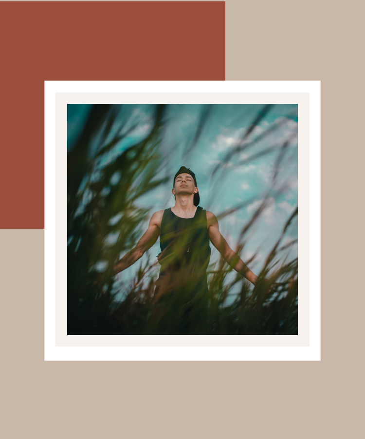 A man standing outdoors among tall grasses with his eyes closed and arms outstretched, enjoying a mindful moment in nature, framed against a terracotta and beige background.