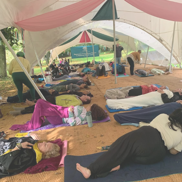 A group of participants lying comfortably on mats and blankets during a guided outdoor breathwork session beneath a spacious canopy, surrounded by trees and natural light.