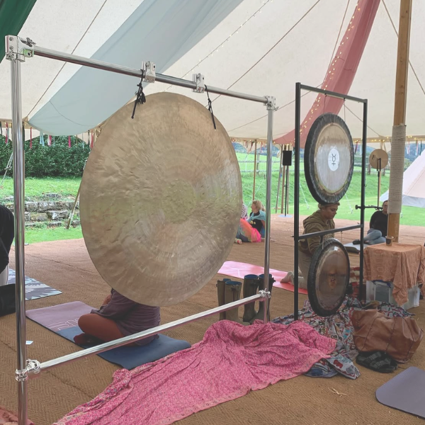 Large suspended gongs and participants resting on yoga mats beneath an outdoor canopy during a peaceful sound healing and relaxation session.