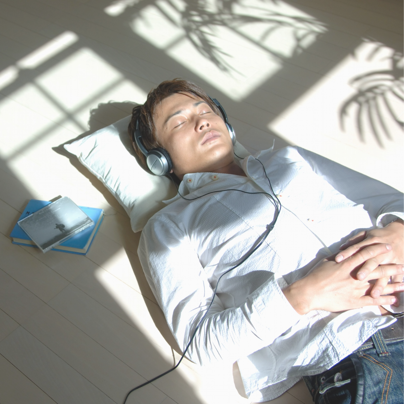 A man lying on a pillow with his eyes closed, wearing headphones and resting his hands on his stomach, surrounded by soft sunlight and leafy shadows in a peaceful indoor setting.