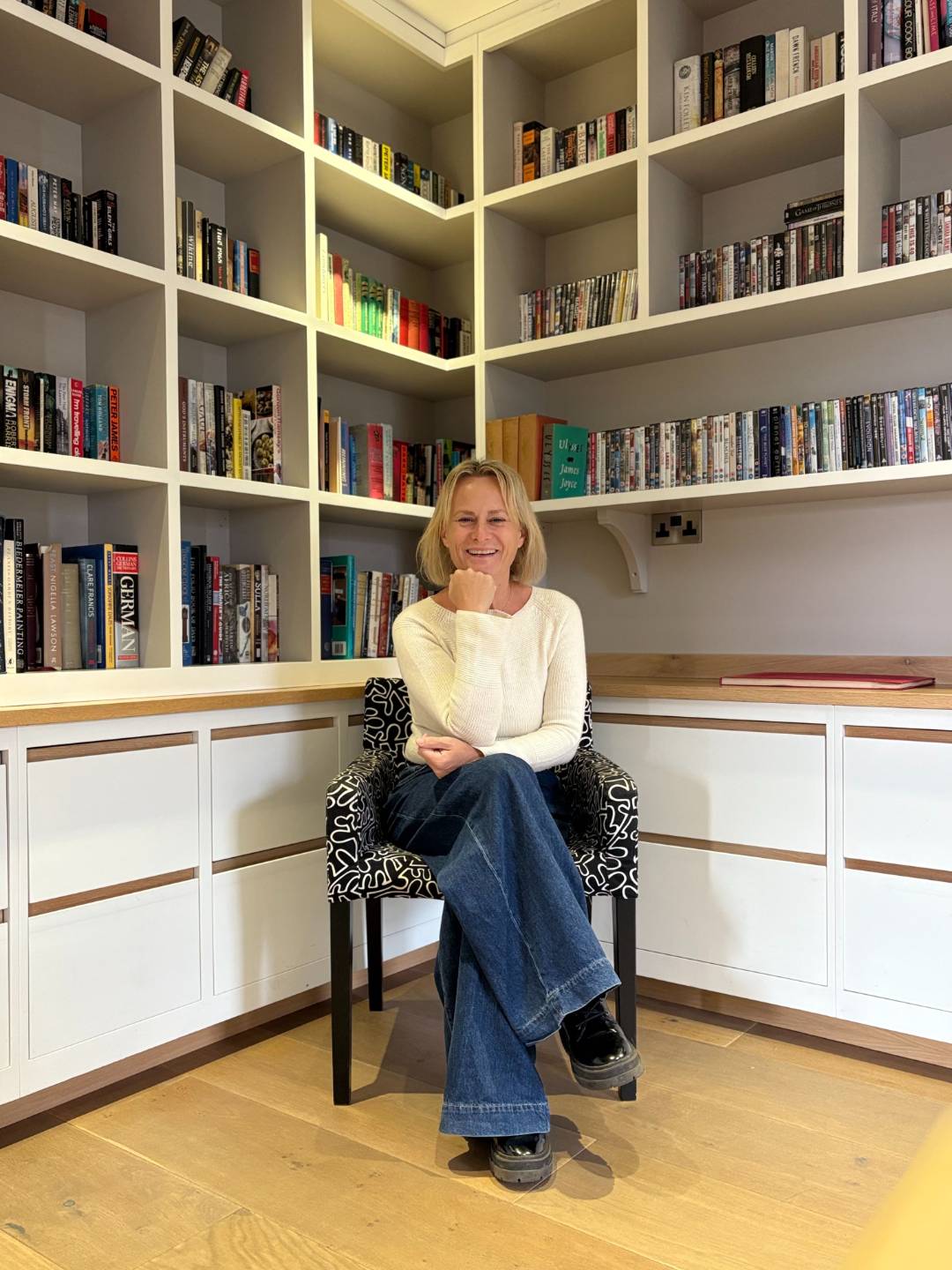 Dee Parker smiling while seated in a patterned armchair, wearing a cream jumper and blue jeans, surrounded by floor-to-ceiling bookshelves in a cosy home library.