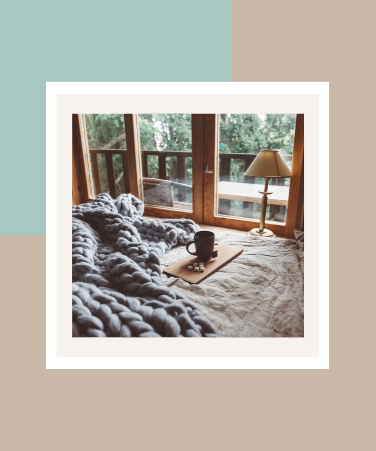 A cosy bedroom with a chunky knitted blanket, a mug on a wooden tray and a bedside lamp overlooking a peaceful woodland view, framed against a soft sage green and beige background.