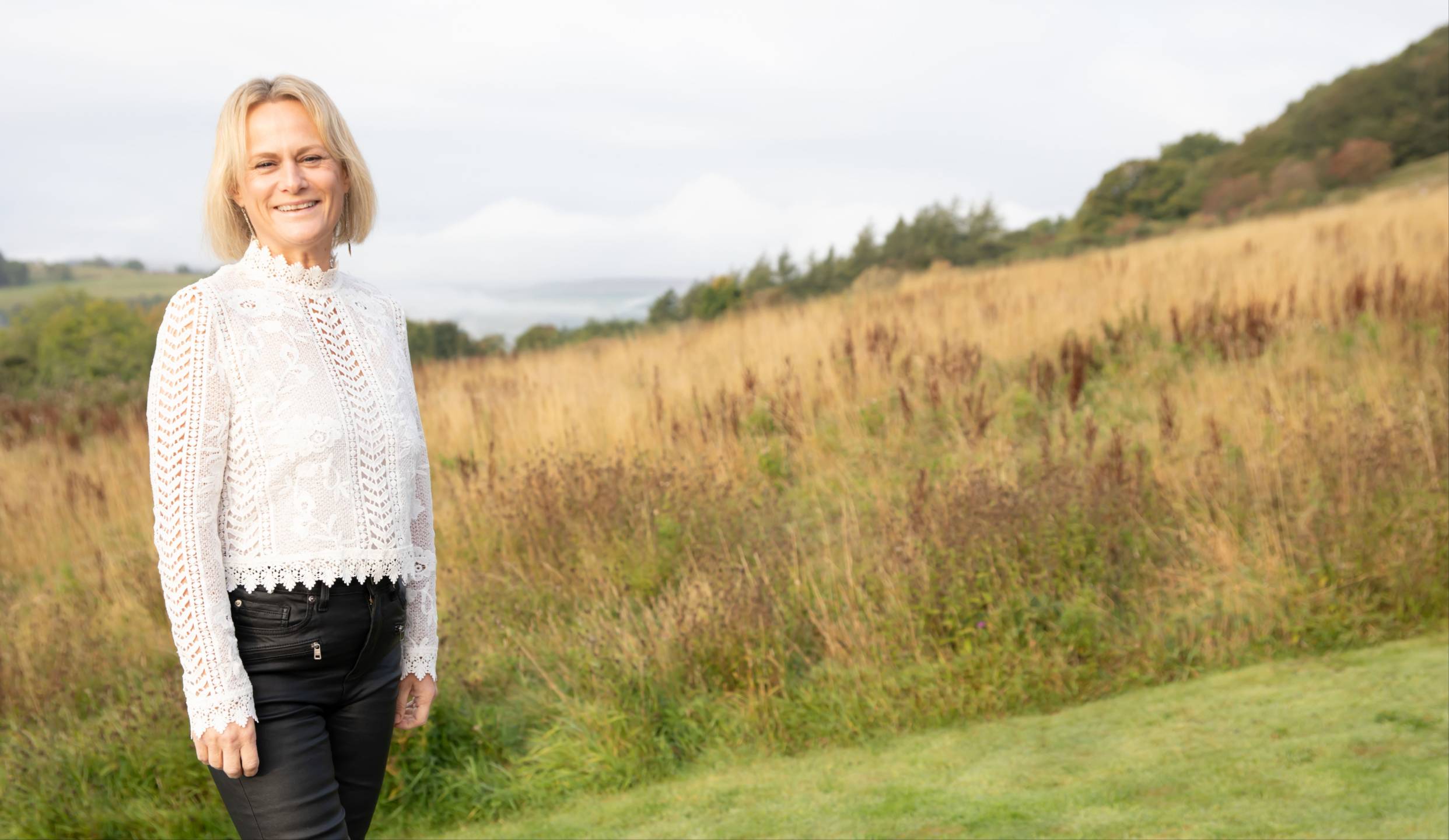 Dee Parker smiling outdoors in a white lace blouse and black trousers, standing in a peaceful countryside setting with golden grass, rolling hills and trees in the background.