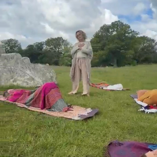 Dee Parker standing barefoot in a grassy field with her hands resting on her chest, guiding an outdoor breathwork session surrounded by nature, blankets and resting participants.