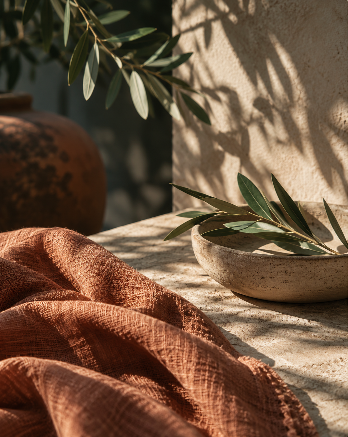 Soft terracotta linen draped across a textured stone surface beside a ceramic bowl of olive branches, with warm sunlight casting delicate leafy shadows on a beige wall.