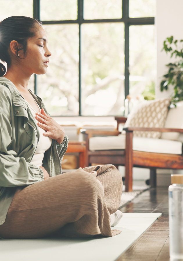 A woman sitting cross-legged on a yoga mat with her eyes closed and one hand resting on her chest, practising mindful breathing in a bright, peaceful room with natural light.