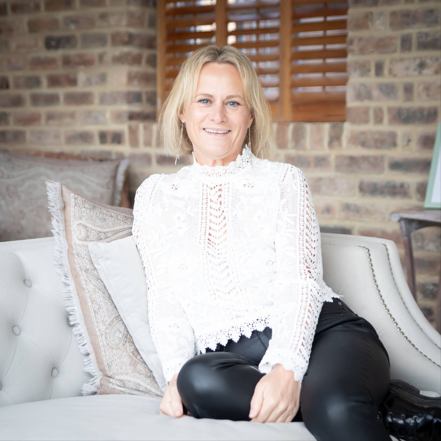 Dee Parker smiling while seated on a light-coloured sofa, wearing a white lace blouse and black trousers, with a rustic brick wall and wooden shutters in the background.