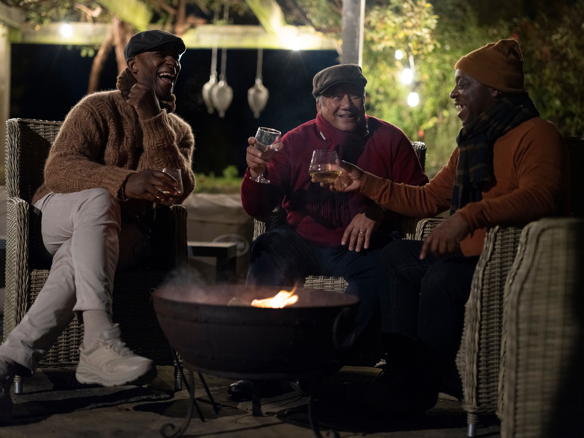 Three friends laughing together around a fire pit at dusk under warm lights