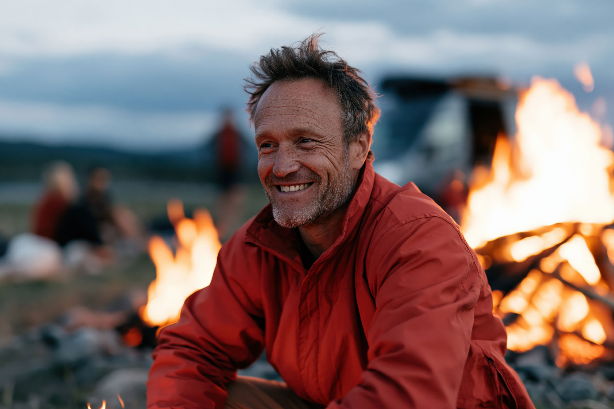 Man in a red jacket laughing beside an open fire