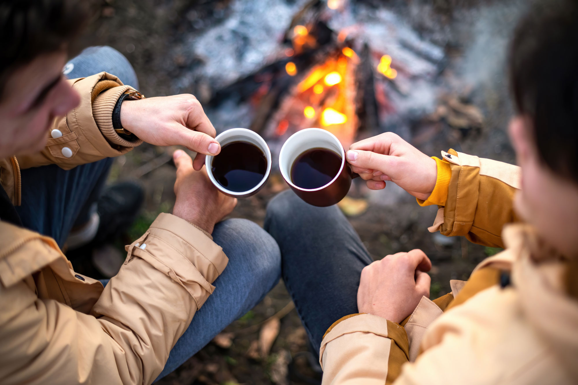 Two enamel mugs raised together beside a campfire