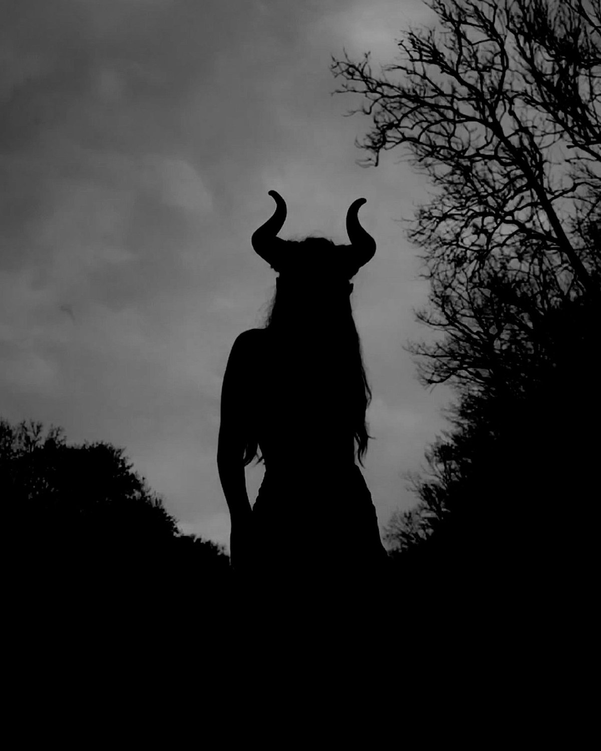 Black-and-white silhouette of Rocío wearing curved horns under a stormy sky, framed by bare trees