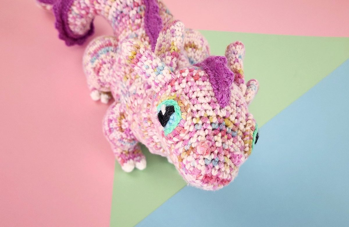 A speckled pink crocheted dragon seen from above on a pastel background