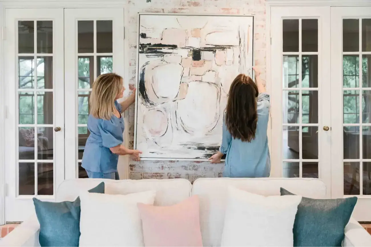 Two home staging professionals positioning large artwork above a sofa to demonstrate proportion, scale, and balance.