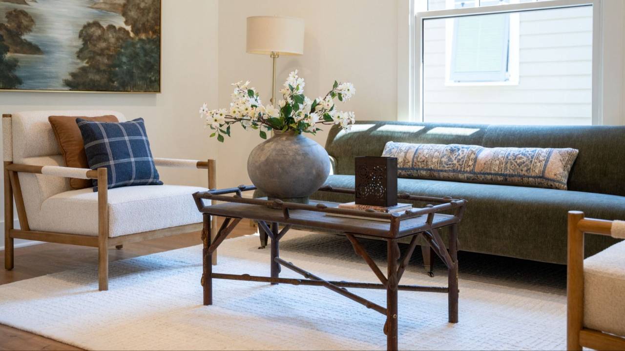 A bright, modern living room featuring a large white fireplace with a textured teal and gold abstract painting resting on the mantle. In front of the fireplace is a dark wood coffee table styled with books, a light blue fluted vase with white flowers, and a bowl of green moss spheres. The seating area includes a plush, cream-colored armchair next to a round wooden side table, and two leather-backed armchairs in the background. Two framed botanical prints hang on the white wall next to a tall, slender potted indoor plant. The room is completed with a light-colored woven area rug and warm hardwood floors.