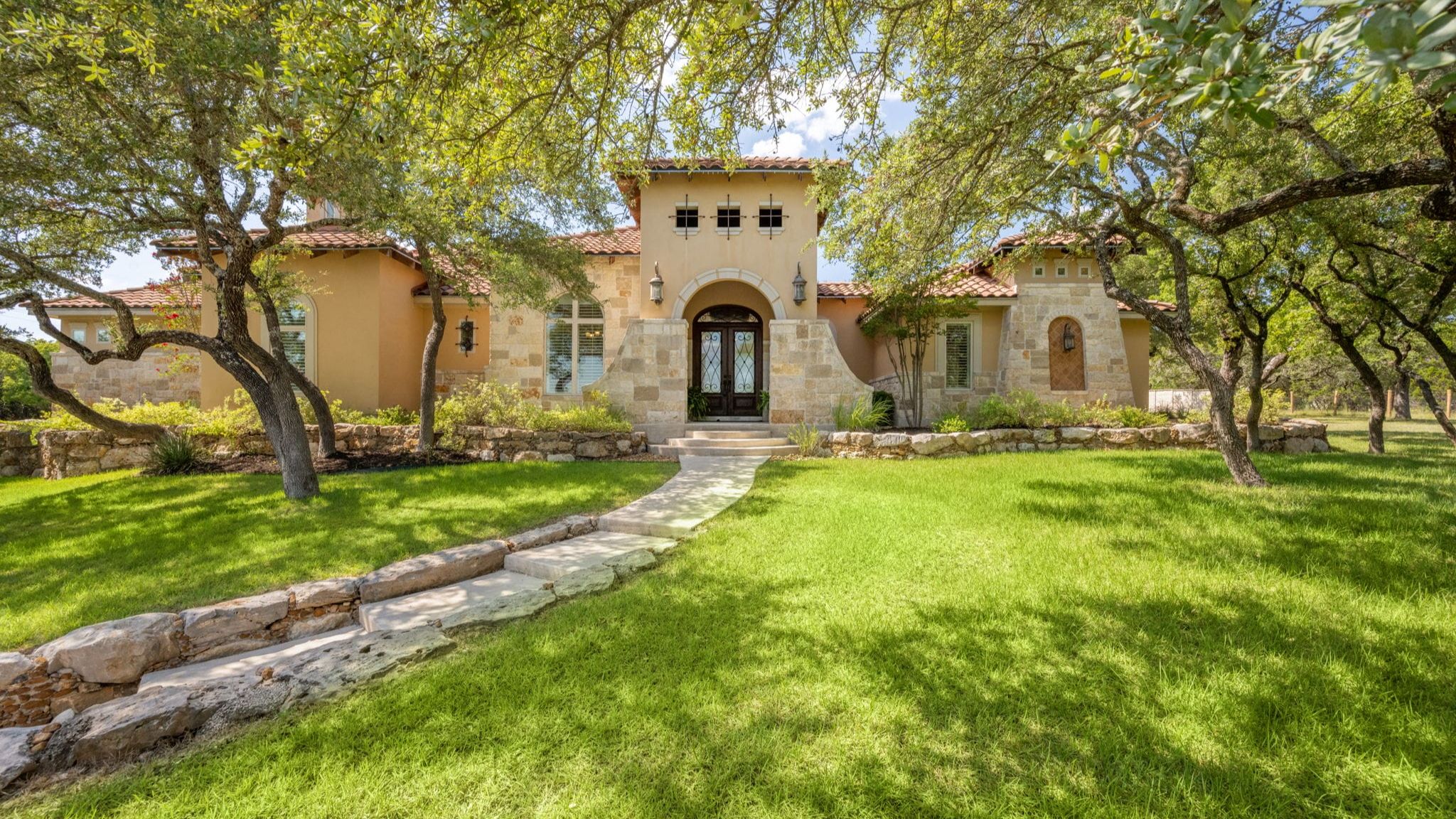 A large, Mediterranean-style luxury house features a light beige stucco exterior combined with light-colored stone accents. The home has a red terracotta tile roof and an arched front entryway with dark double doors. A stone walkway cuts through a lush, vibrant green lawn leading up to the house, shaded by mature, leafy oak trees.