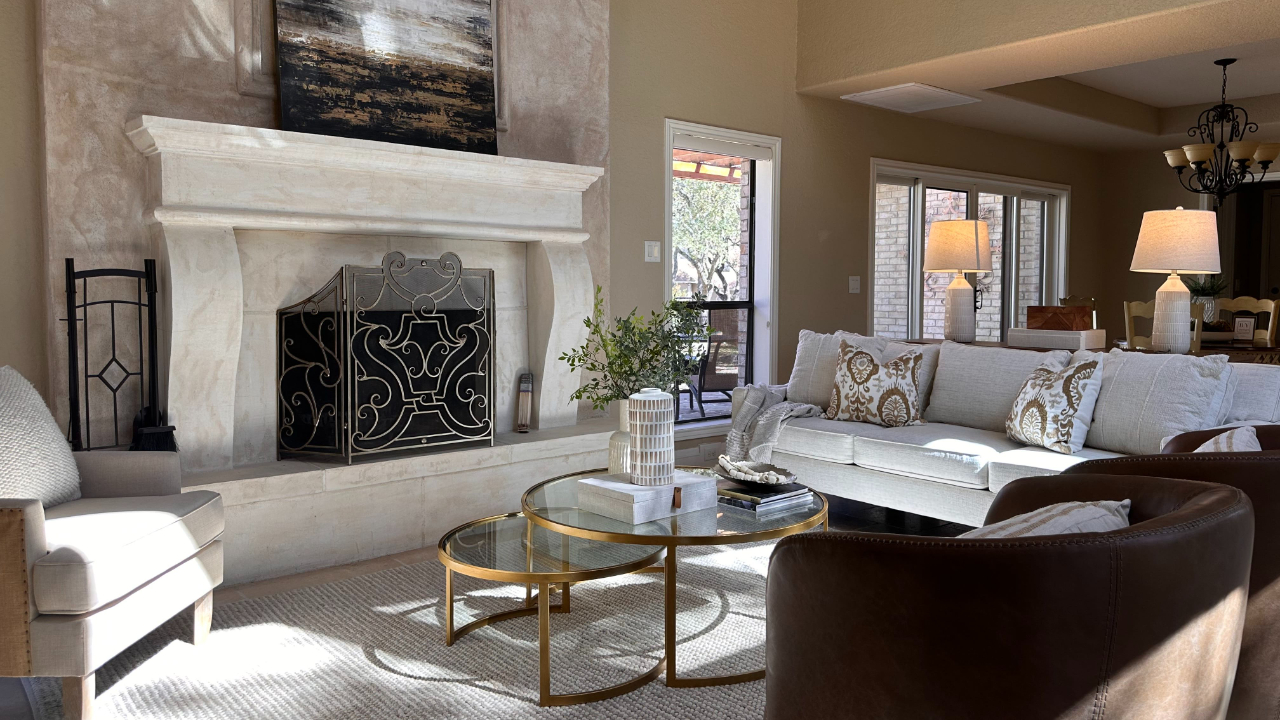 A bright, spacious living room with high ceilings, featuring a large stone fireplace with an ornate screen, a white sofa with patterned accent pillows, a round glass nesting coffee table, and comfortable armchairs arranged over a light textured area rug.