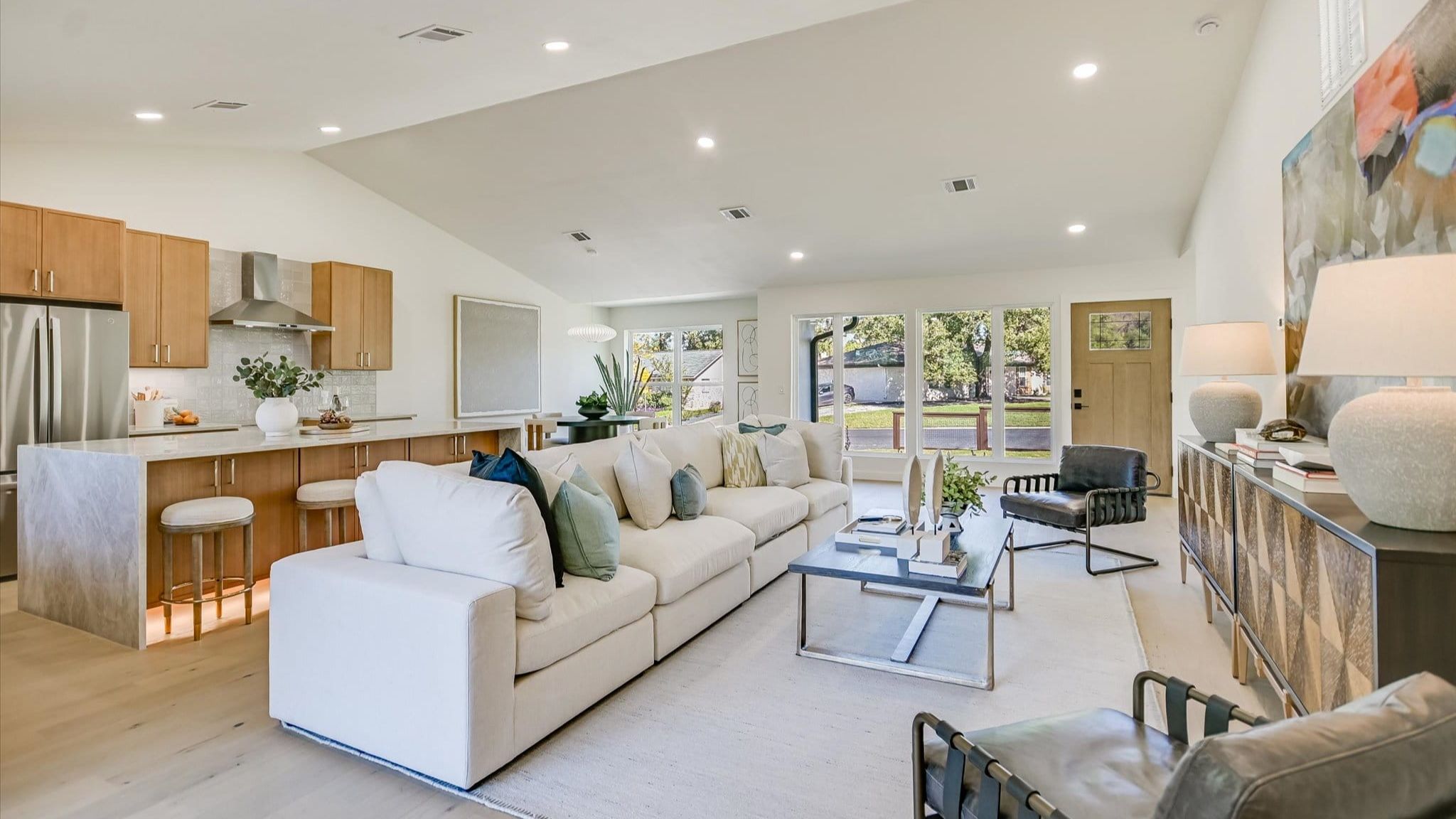 An open-concept living room and kitchen featuring bright, modern design with vaulted ceilings and recessed lighting. A long, cream-colored sectional sofa with accent pillows sits on a light area rug, facing a dark metal coffee table and two black leather accent chairs. In the background, the adjacent kitchen shows light wood cabinetry, a marble-waterfall island with barstools, and stainless steel appliances. Large windows and an open wooden front door bring natural light into the space.