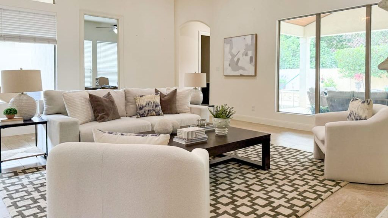 Bright contemporary living room with neutral staging, featuring a cream sofa, curved accent chairs, patterned area rug, dark wood coffee table, and large windows overlooking an outdoor patio.