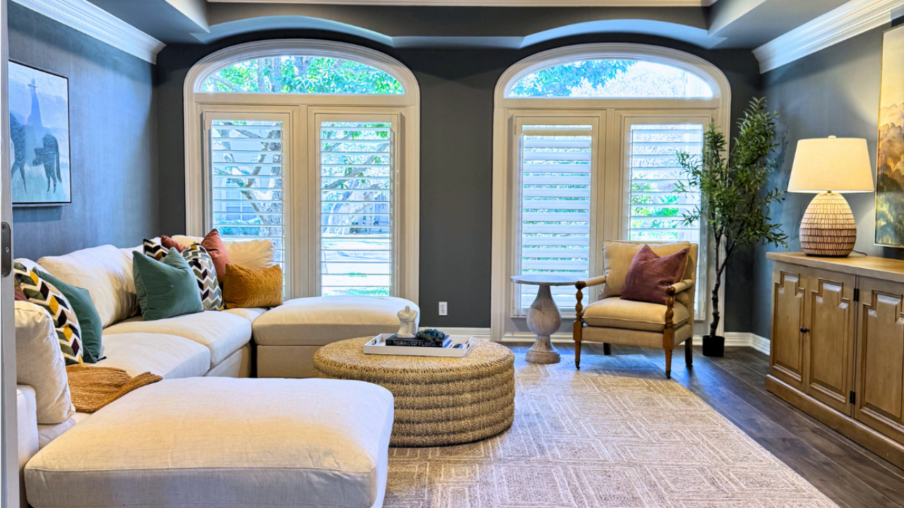 A luxurious dining room with taupe walls, featuring a long dark wood table surrounded by unique, cream-colored curved bouclé dining chairs on a large patterned area rug, lit by two crystal chandeliers.