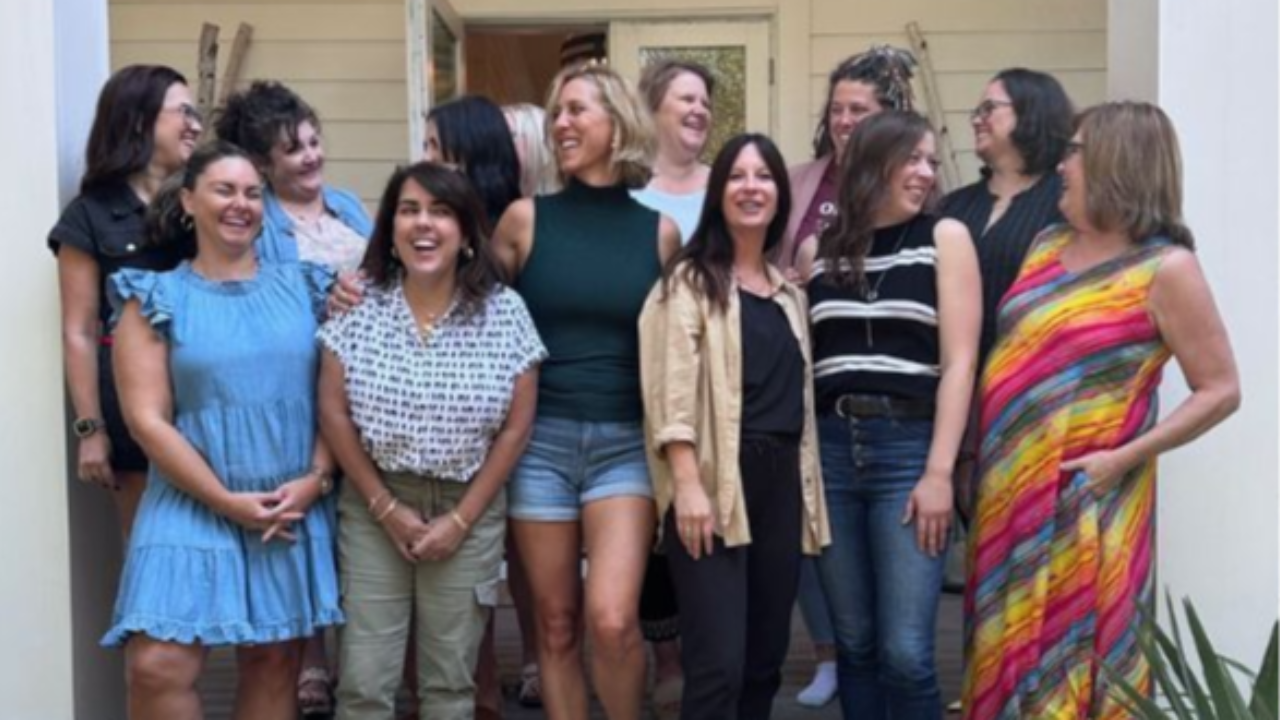 A group of women standing on a porch, smiling, laughing, and looking at each other in a casual, friendly moment.