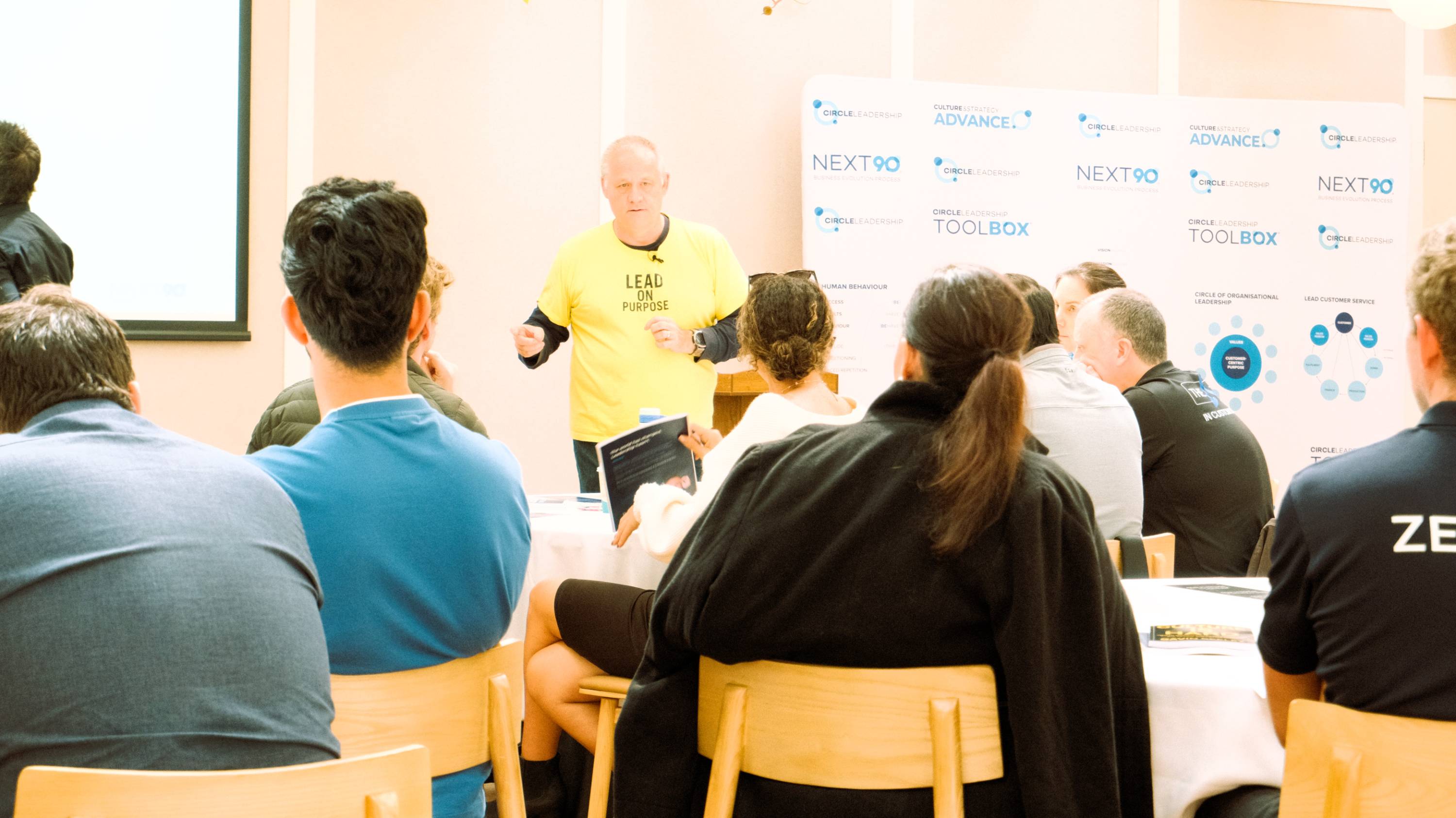 A facilitator leads a Circle Leadership workshop with participants seated around tables.