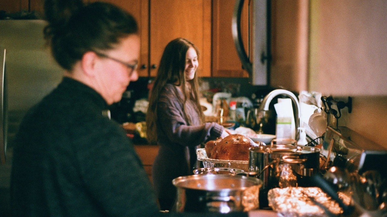 Two women cooking a holiday meal together in a warm kitchen, one smiling as she helps with the turkey.