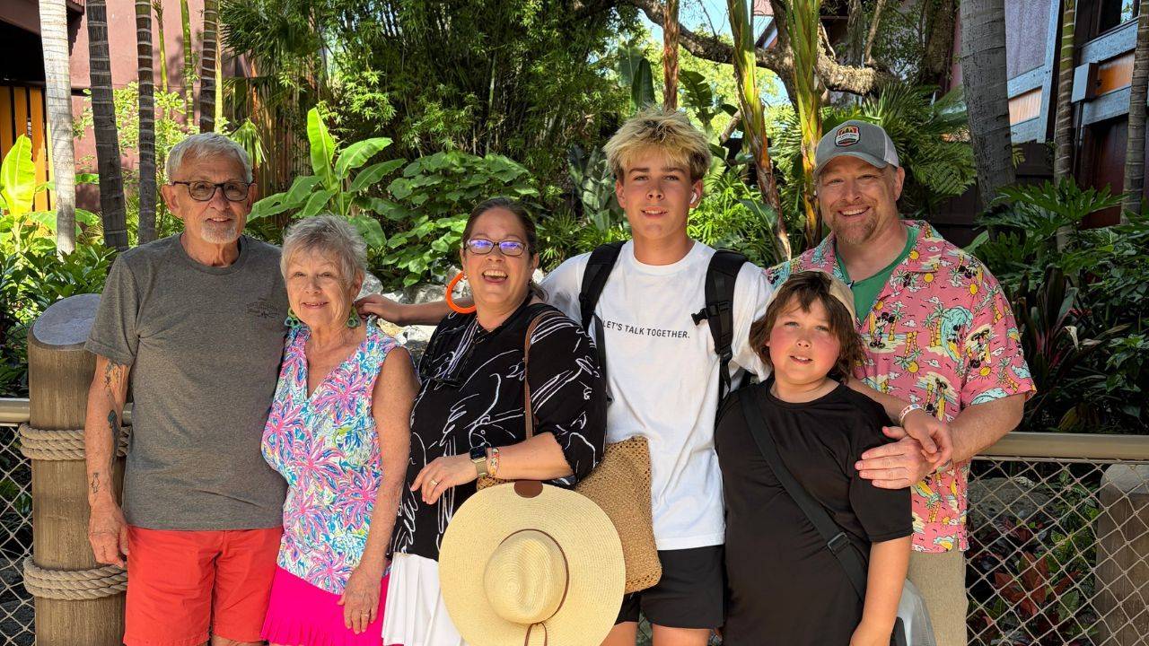 Six family members, including two older adults, two adults, a teenage boy, and a younger boy, pose together outdoors surrounded by lush tropical greenery.