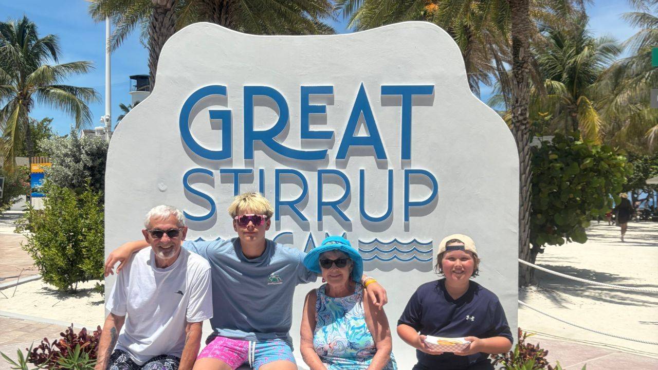 Two older adults and two boys sit together in front of a large Great Stirrup Cay sign, with palm trees and a sunny beach setting behind them.