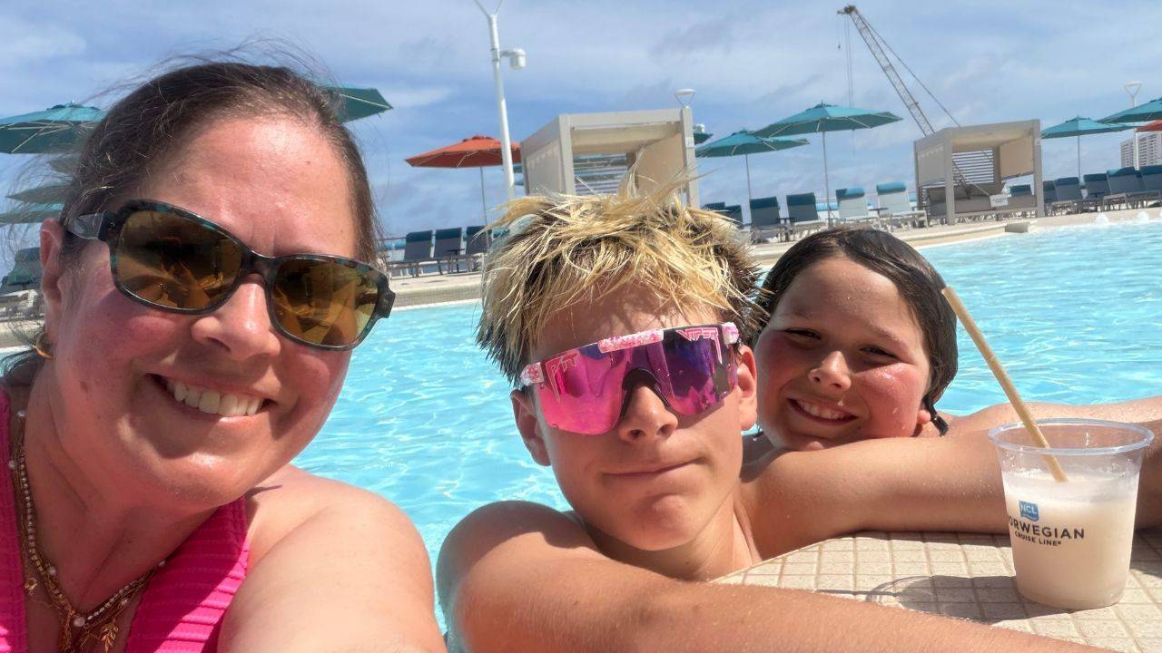 A woman and two boys smile together beside a cruise ship swimming pool on a sunny day. The teenage boy wears bright pink sunglasses, and a tropical drink sits on the pool ledge.