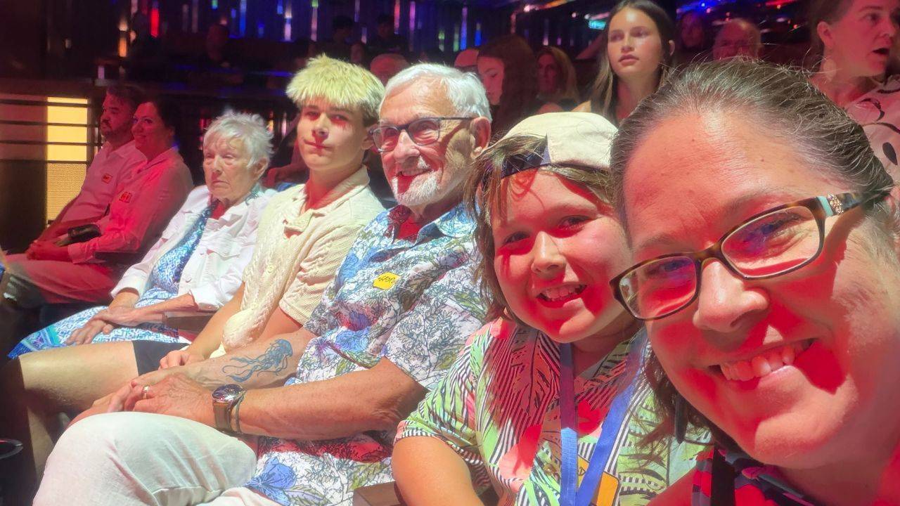Five family members sit together in a theater or entertainment venue under colorful red and blue lighting, smiling toward the camera before a show.