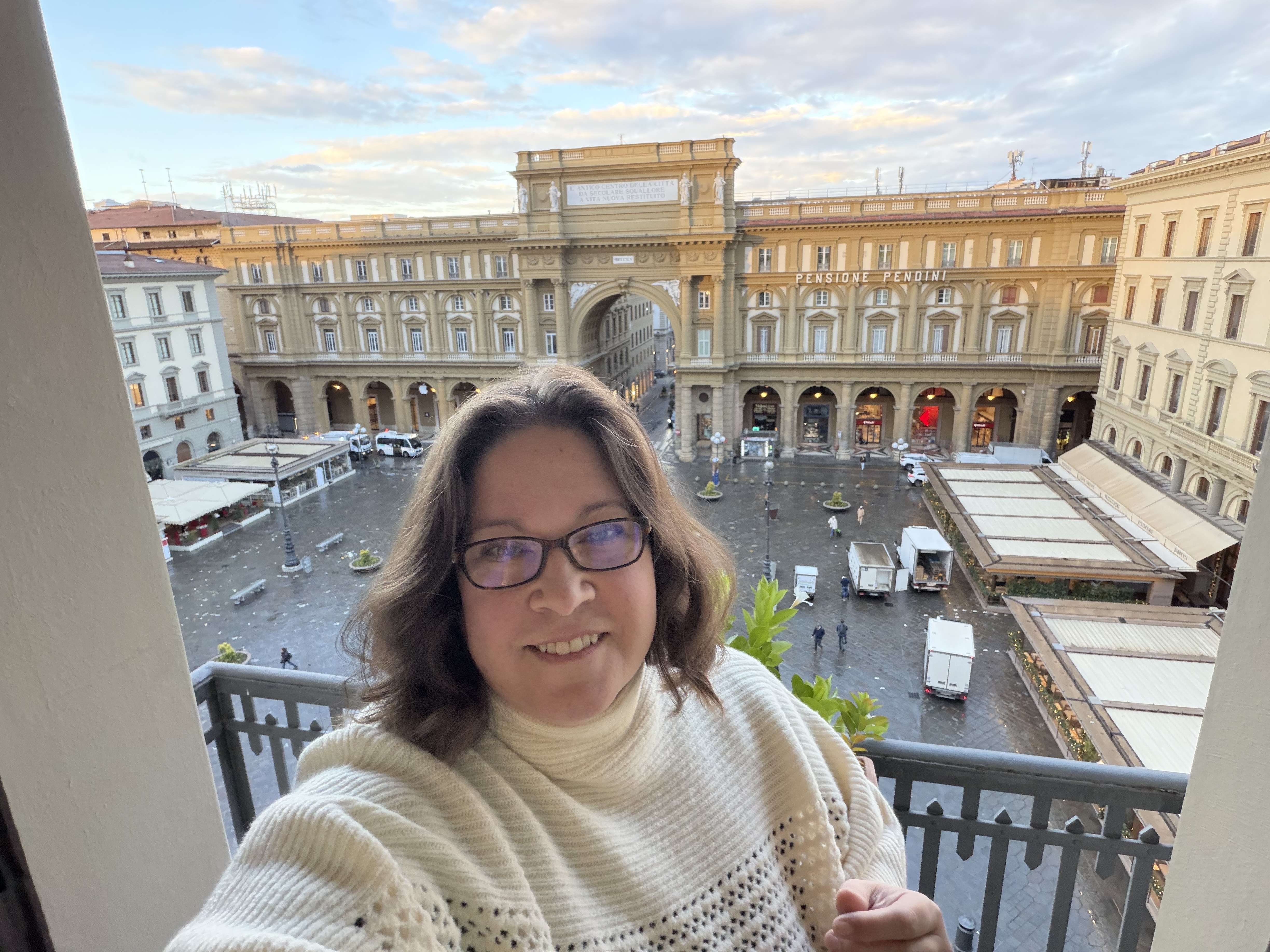 Tamsen Horton smiles on a balcony overlooking the Piazza della Repubblica in Florence, Italy, wearing a white knit sweater with the historic Arcone di Piazza in the background at sunset.